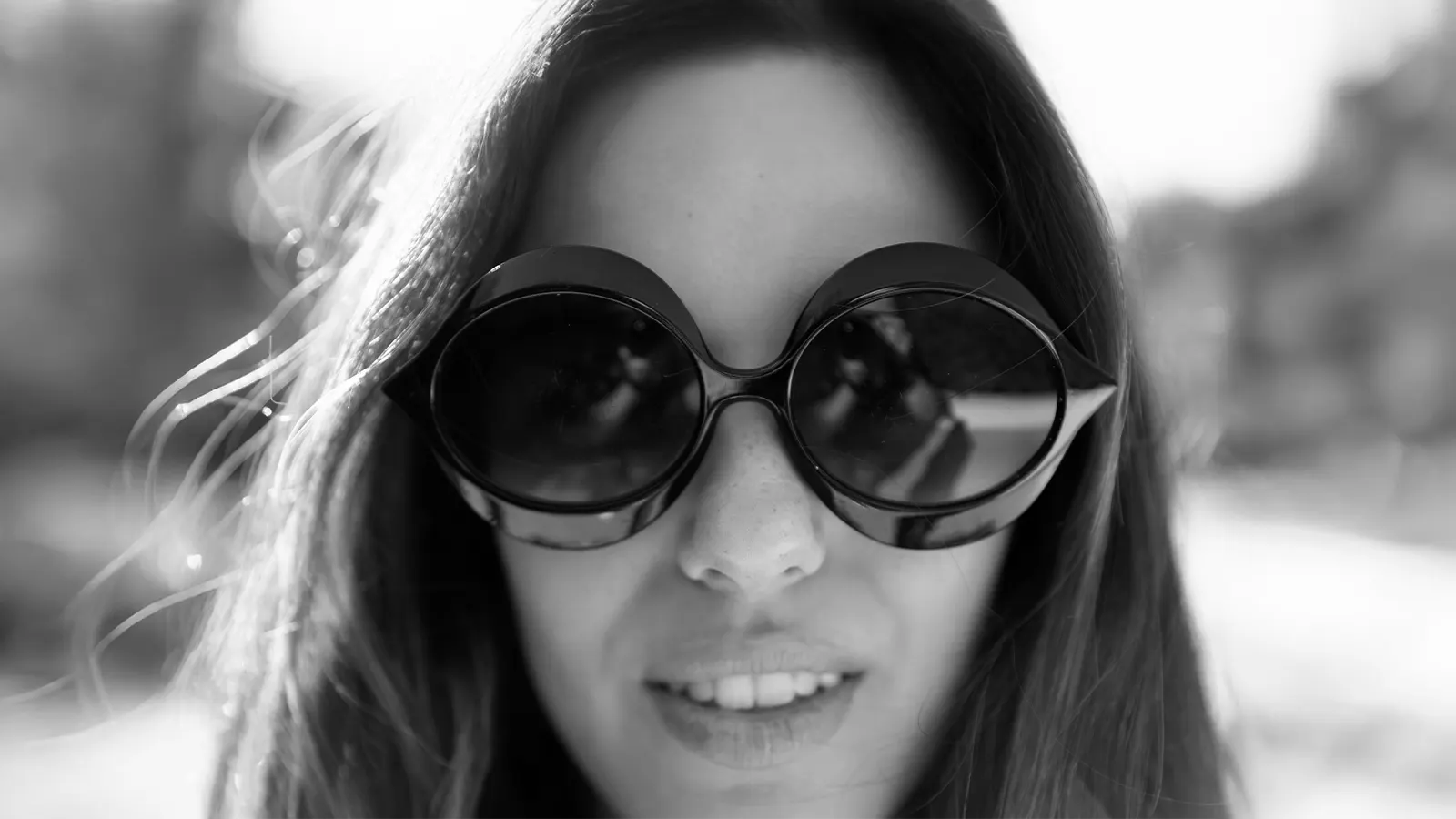 This black and white close-up features a woman wearing very large, dark, round sunglasses. The unique, almost goggle-like design with a thick frame around the lens is a distinct, dramatic style, evoking a bold fashion statement.