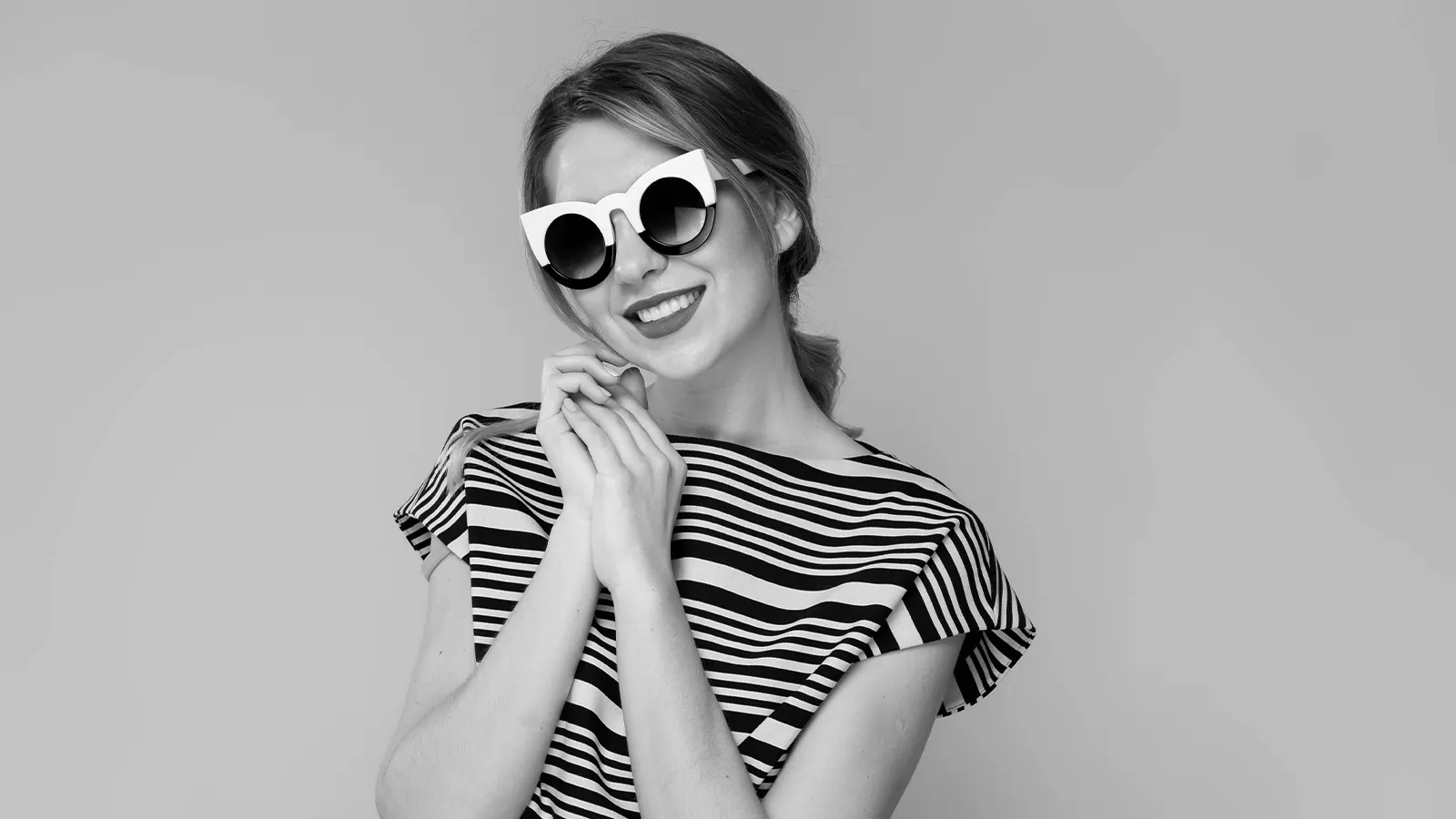 This black and white image shows a smiling woman in a striped top and red lipstick. She is wearing bold, oversized sunglasses featuring a thick, contrasting white and black frame that makes a strong, graphic fashion statement.