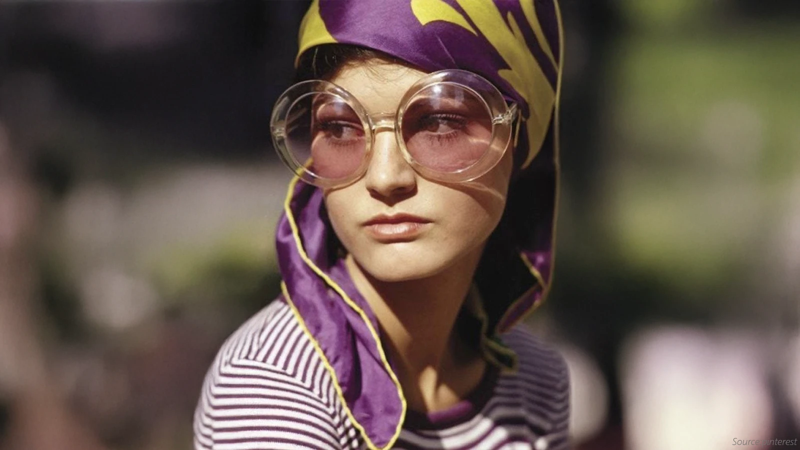 This portrait shows a person wearing a striped shirt and a colourful purple and yellow headscarf. They are wearing very large, round sunglasses with thick, light-colored, clear frames and pink-tinted lenses, characteristic of 1960s and 1970s fashion.
