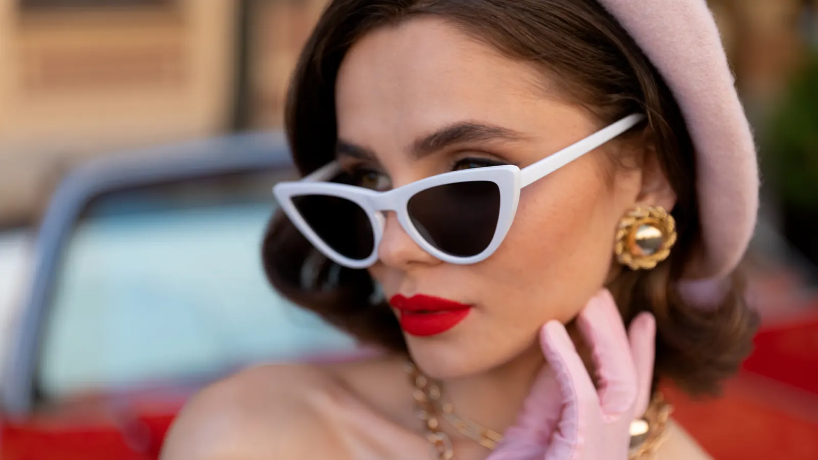 This vibrant portrait shows a woman in a retro style with bright red lipstick and pink gloves. She is wearing bold, white-framed cat-eye sunglasses, large gold earrings, and a chunky gold necklace, epitomising mid-century glamour.