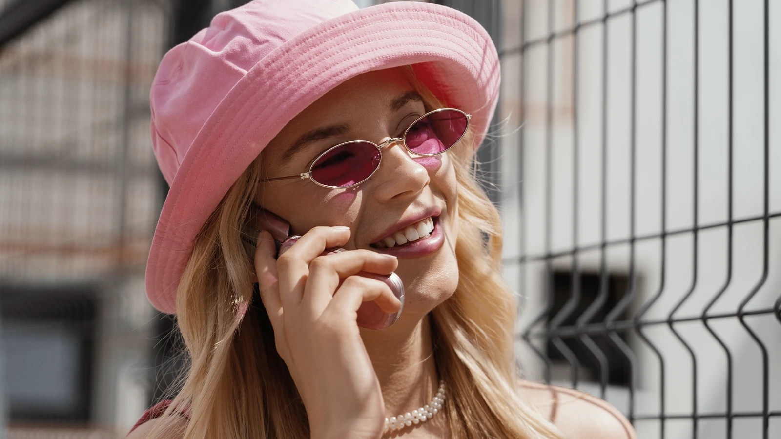 A smiling woman is talking on the phone outdoors, wearing stylish pink-tinted oval sunglasses that match her pink bucket hat. Her blonde hair, pearl necklace, and relaxed vibe give the image a trendy, summery feel.