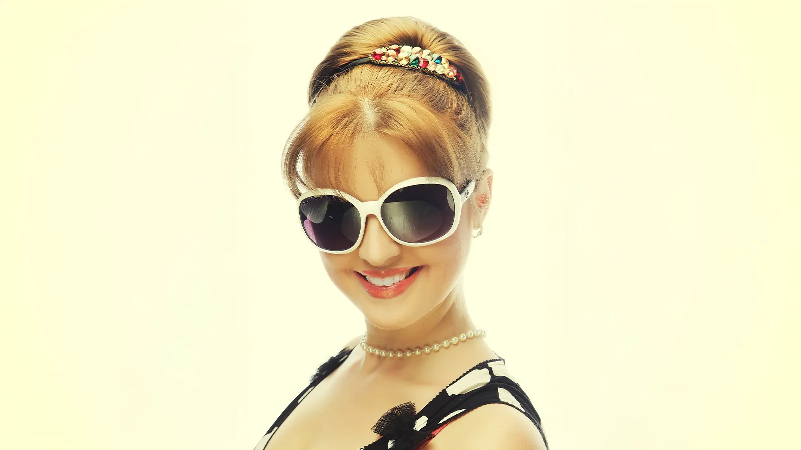 The image features a woman in a retro-inspired look with a styled updo and a pearl headband. She is wearing white-framed, oversized sunglasses and a pearl necklace, smiling brightly against a pale yellow background.