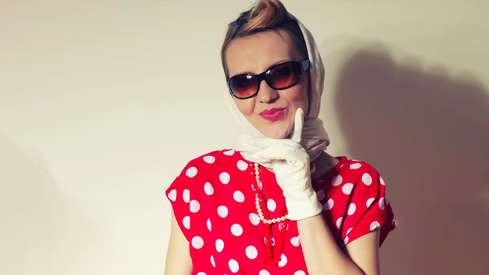This vibrant image features a woman in a retro style, wearing a red polka-dot top, pearls, and a headscarf. She is posing with white gloves and dark, oval-shaped wrap-around sunglasses, completing the dramatic vintage look.