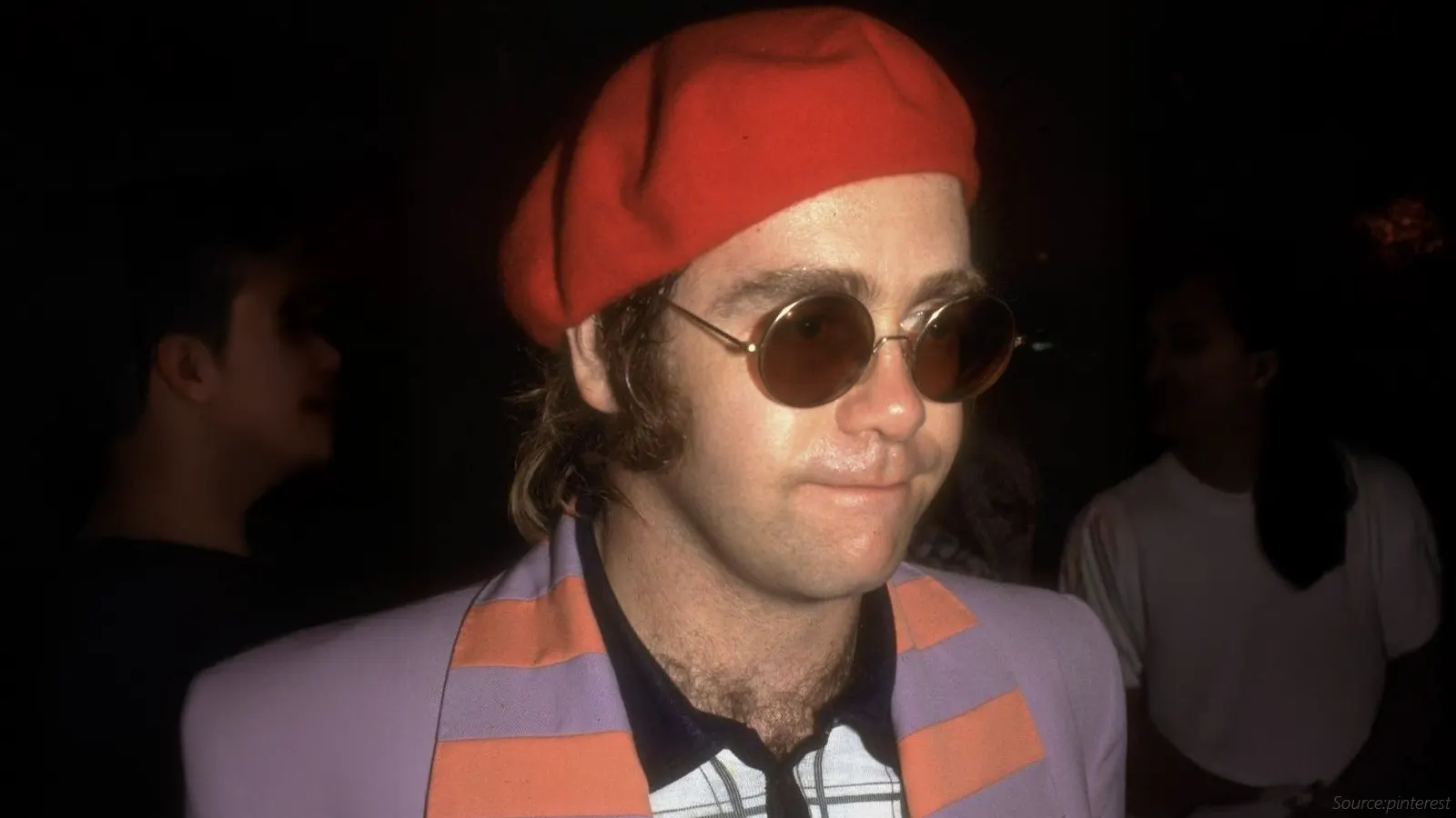 This portrait features a man in a red beret and a lavender jacket with orange stripes. He is wearing small, round sunglasses with thin metal frames and brown-tinted lenses, a style popularised in the 1970s.