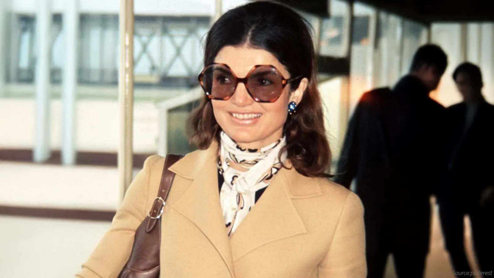 This iconic image features a woman in a camel-colored coat and a patterned silk scarf. She is wearing extremely large, hexagonal tortoiseshell sunglasses, a distinctively glamorous and oversized eyewear style popular in the 1970s.