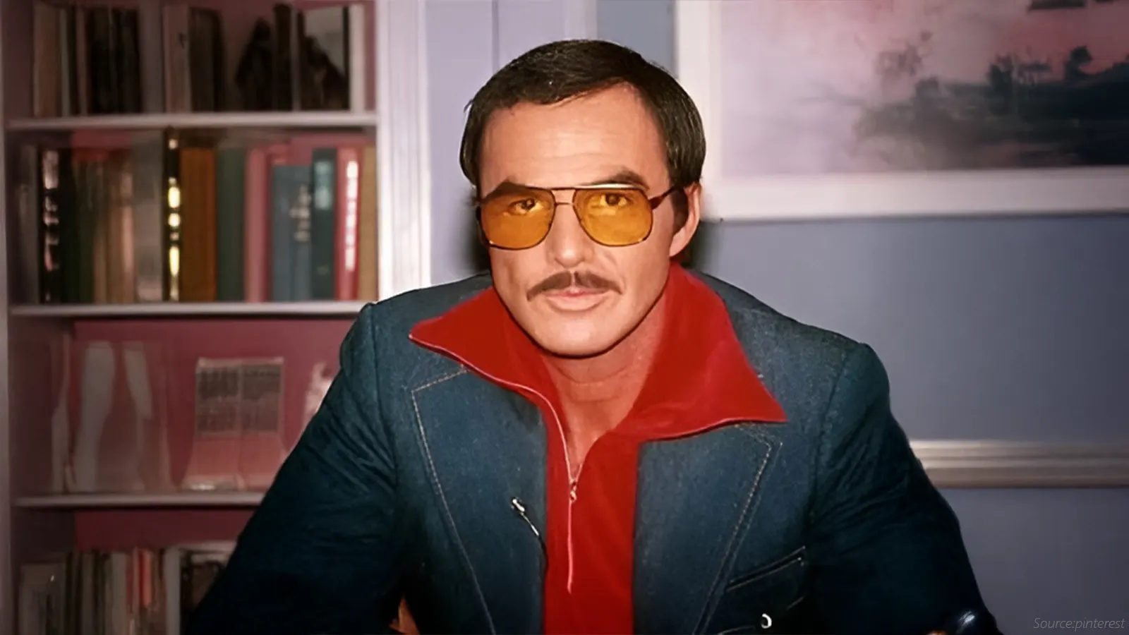 This image features a man with a moustache, wearing a denim jacket and a vibrant red collared shirt. He is wearing classic aviator-style sunglasses with a double brow bar, featuring bold orange or yellow-tinted lenses, a popular trend of the 1970s.