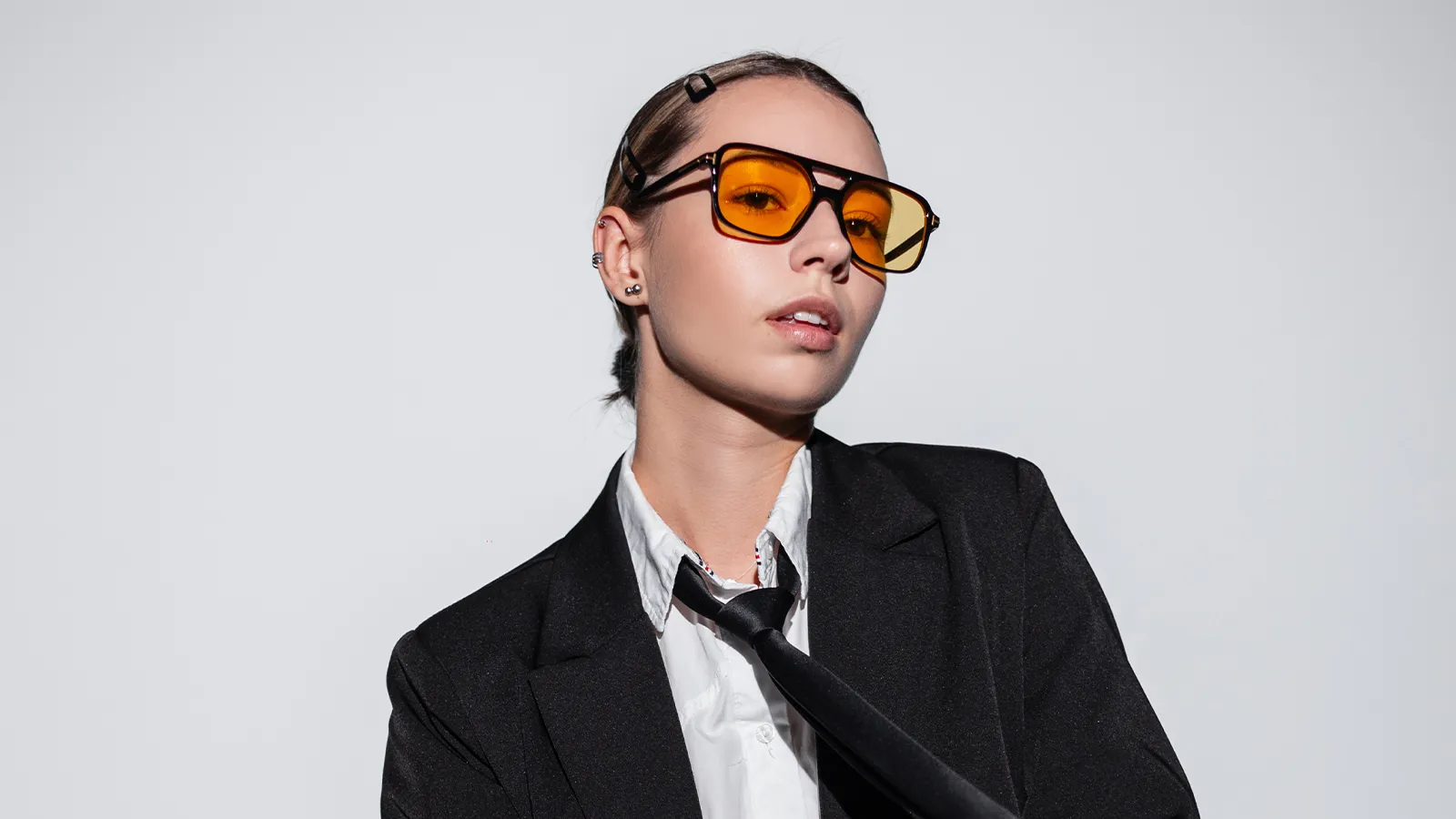 This image shows a person in a bold Y2K-inspired style. The girl is wearing a black suit jacket, a white shirt, and a loose black tie. The look is completed with large, rectangular sunglasses with orange-tinted lenses and minimalist hair clips.