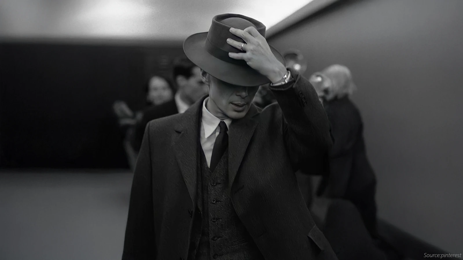 Cillian Murphy’s Oppenheimer in a vintage three-piece suit and tie, adjusting his wide-brimmed hat, captured in a classic black-and-white style with a dramatic, noir-inspired look.
