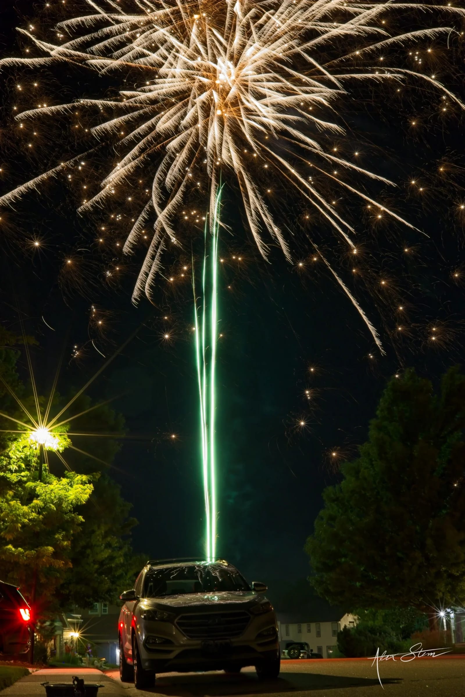 Vibrant fireworks burst in the night sky, illuminating a parked car in the foreground against a backdrop of trees and buildings.