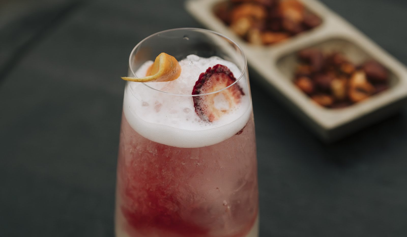 A strawberry cocktail with an orange curl on an OKU napkin, nuts alongside.