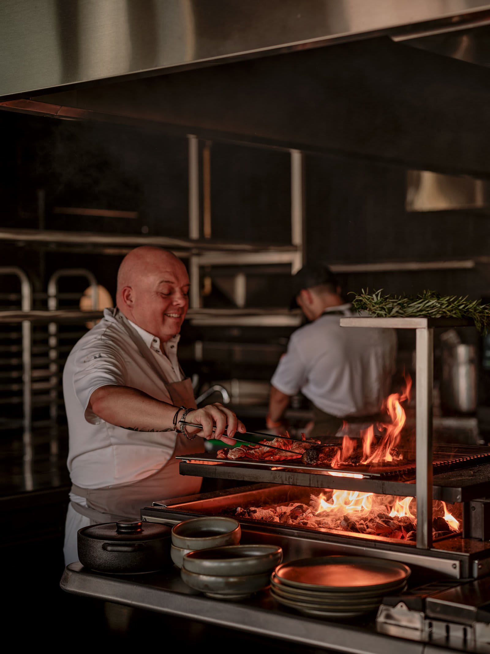 A chef grilling over open flame, smiling, a second cook behind him.