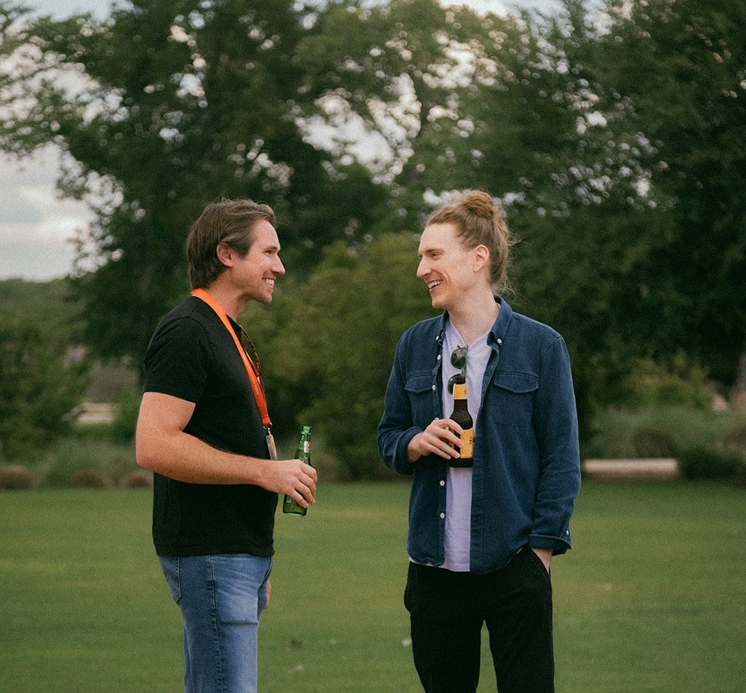 Two attendees laughing together on the lawn, drinks in hand.