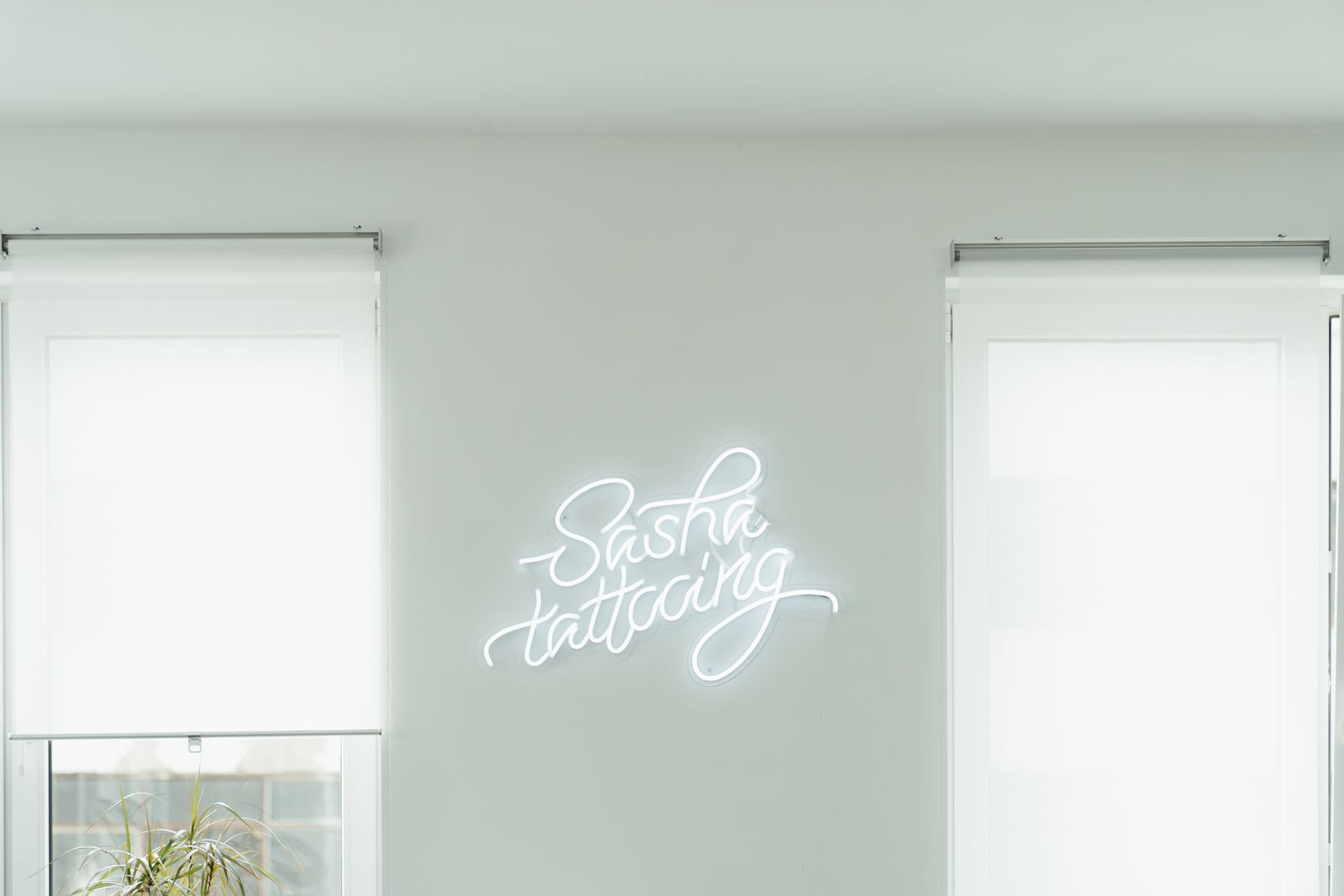 White cursive 'Sasha tattooing' neon script glowing on a pale wall between two bright windows