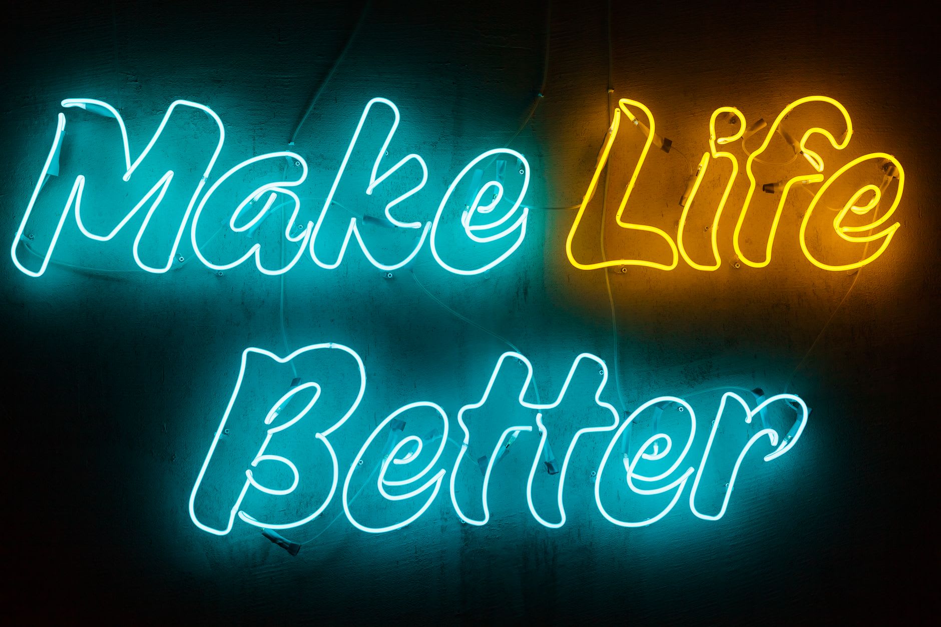 "Make Life Better" script neon on a wall, "Make" and "Better" in teal with "Life" in glowing yellow.