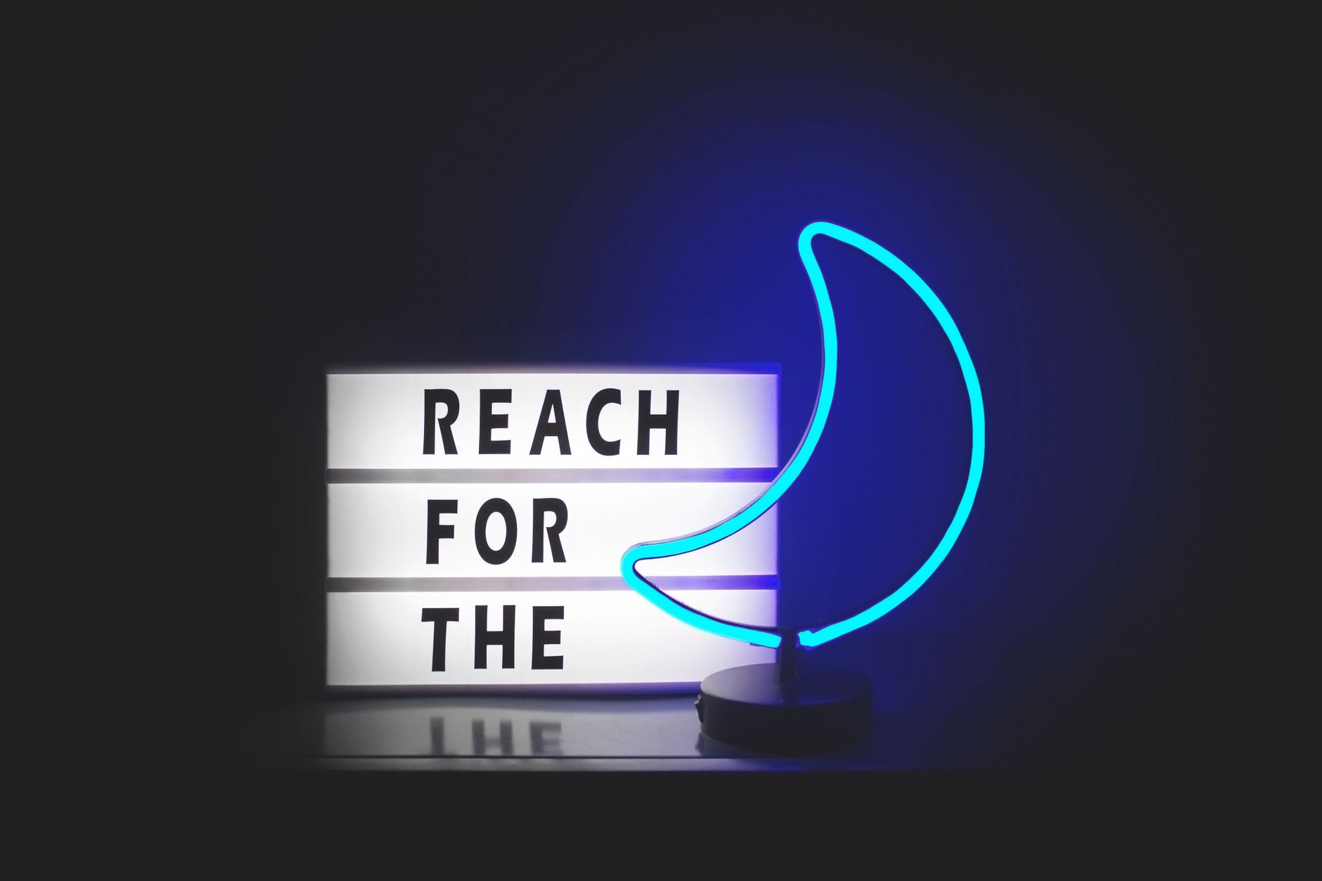 Blue crescent moon neon lamp glowing beside a lightbox reading "Reach for the" against a black background.