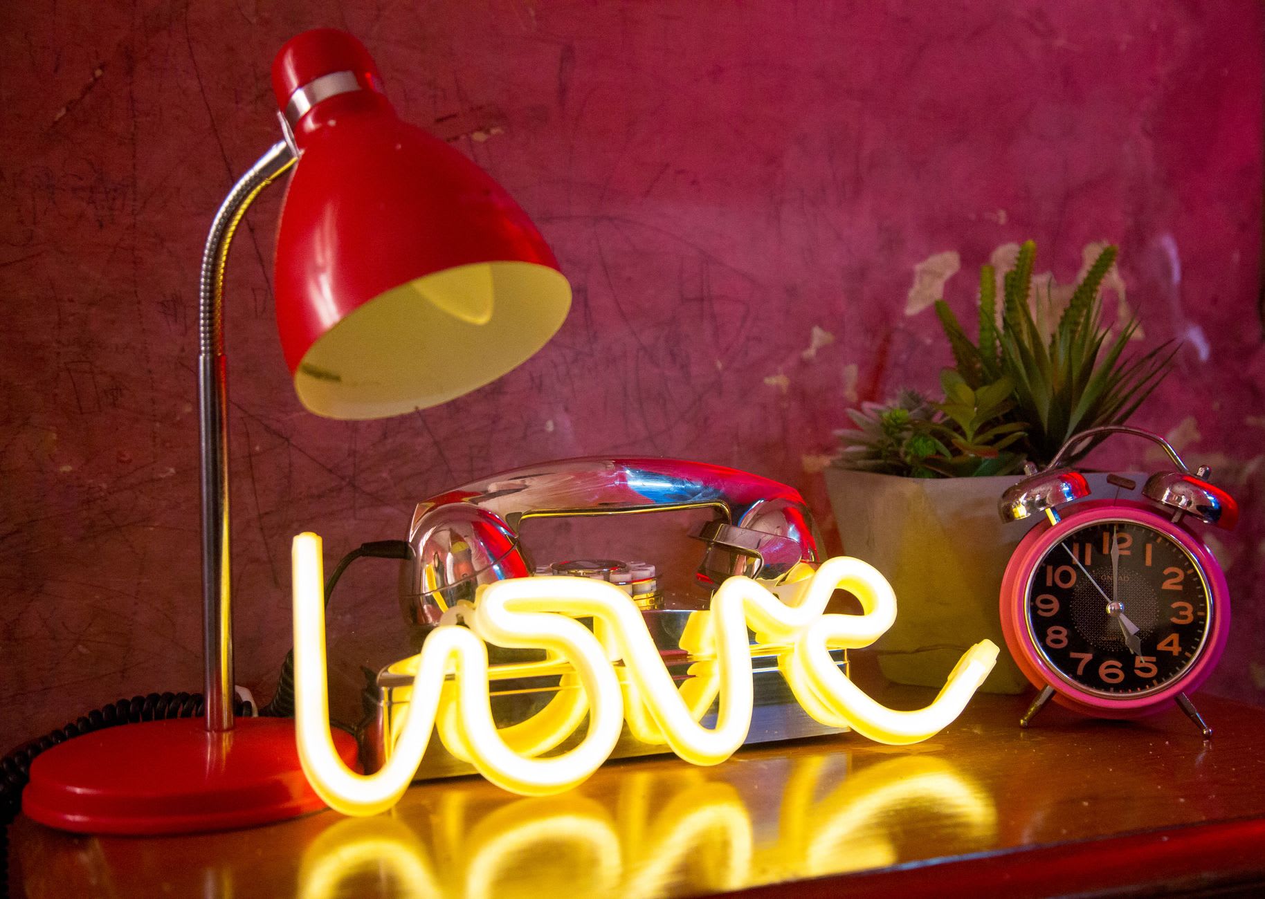 A glowing yellow 'love' neon sign on a wooden table with a red lamp, chrome phone and pink clock against a pink wall.