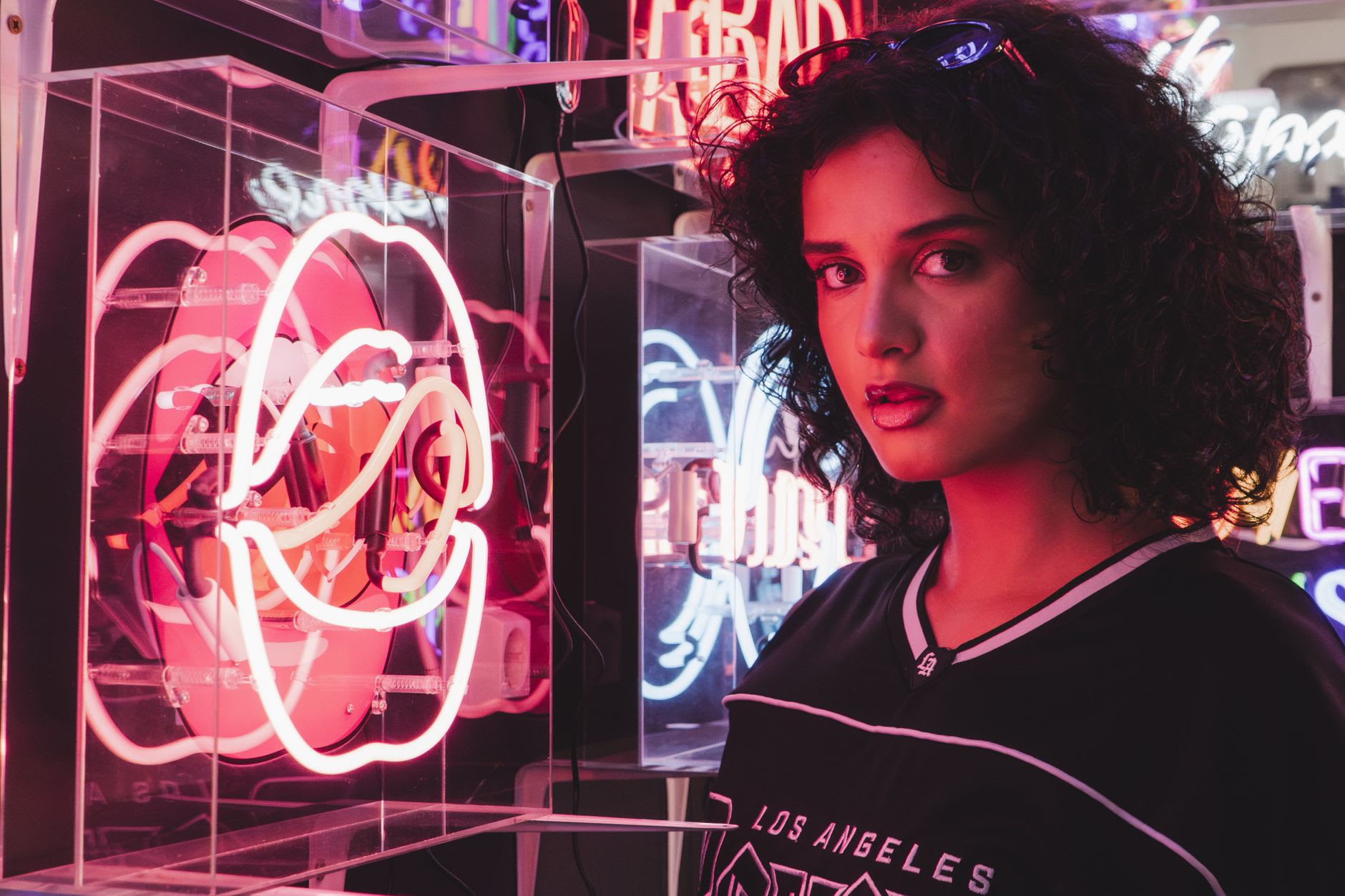 A woman stands confidently among colorful Los Angeles neon signs in a vibrant, glowing space.