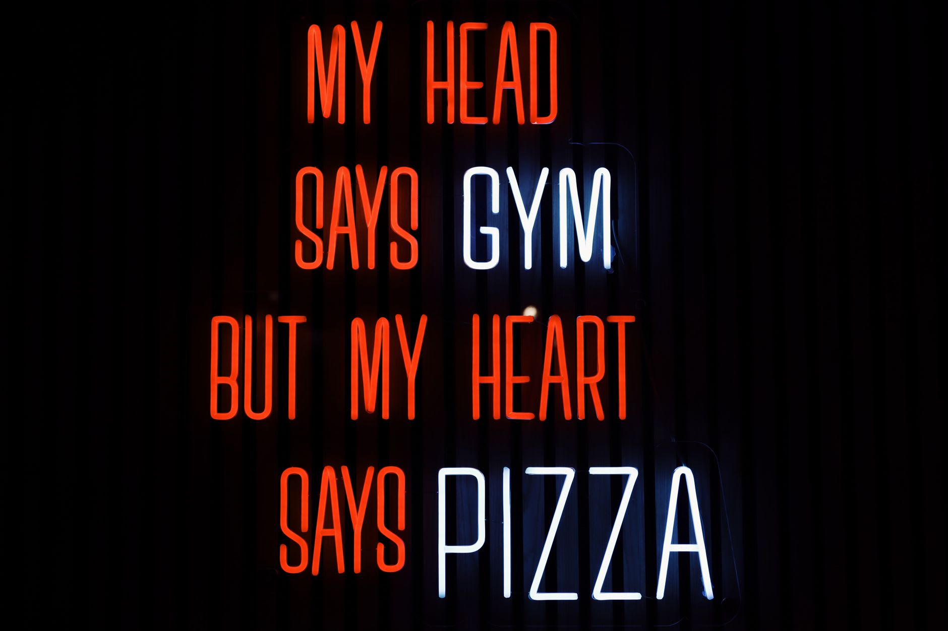 Red and white neon sign reading 'My head says gym but my heart says pizza' glowing on a dark wall.