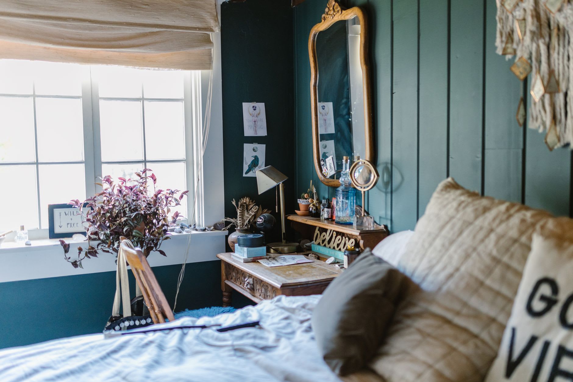 A cozy boho bedroom with deep teal walls, a vintage ornate mirror, crystals, plants and a glowing 'believe' sign.