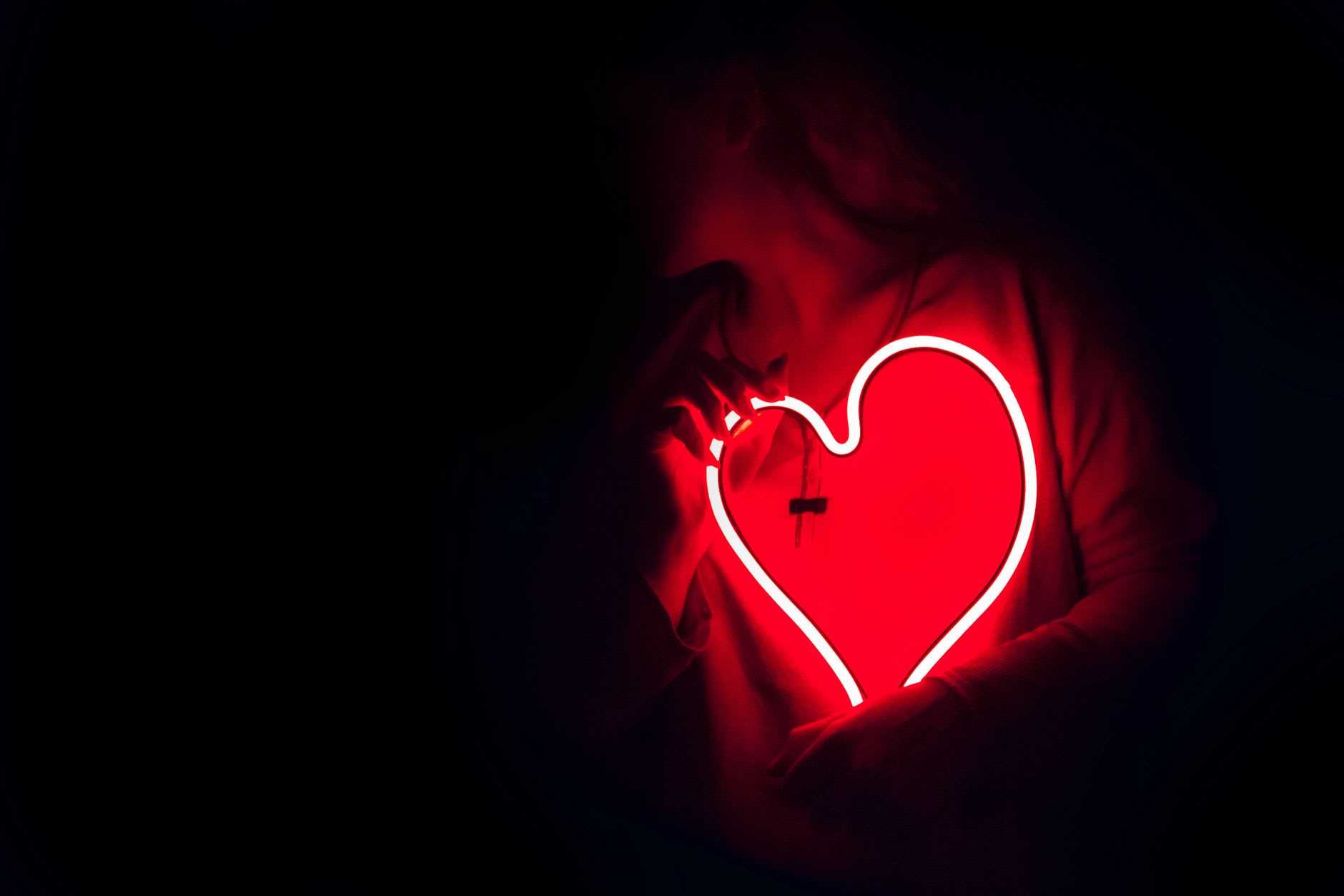 A person's silhouette cradles a glowing red neon heart in the dark.