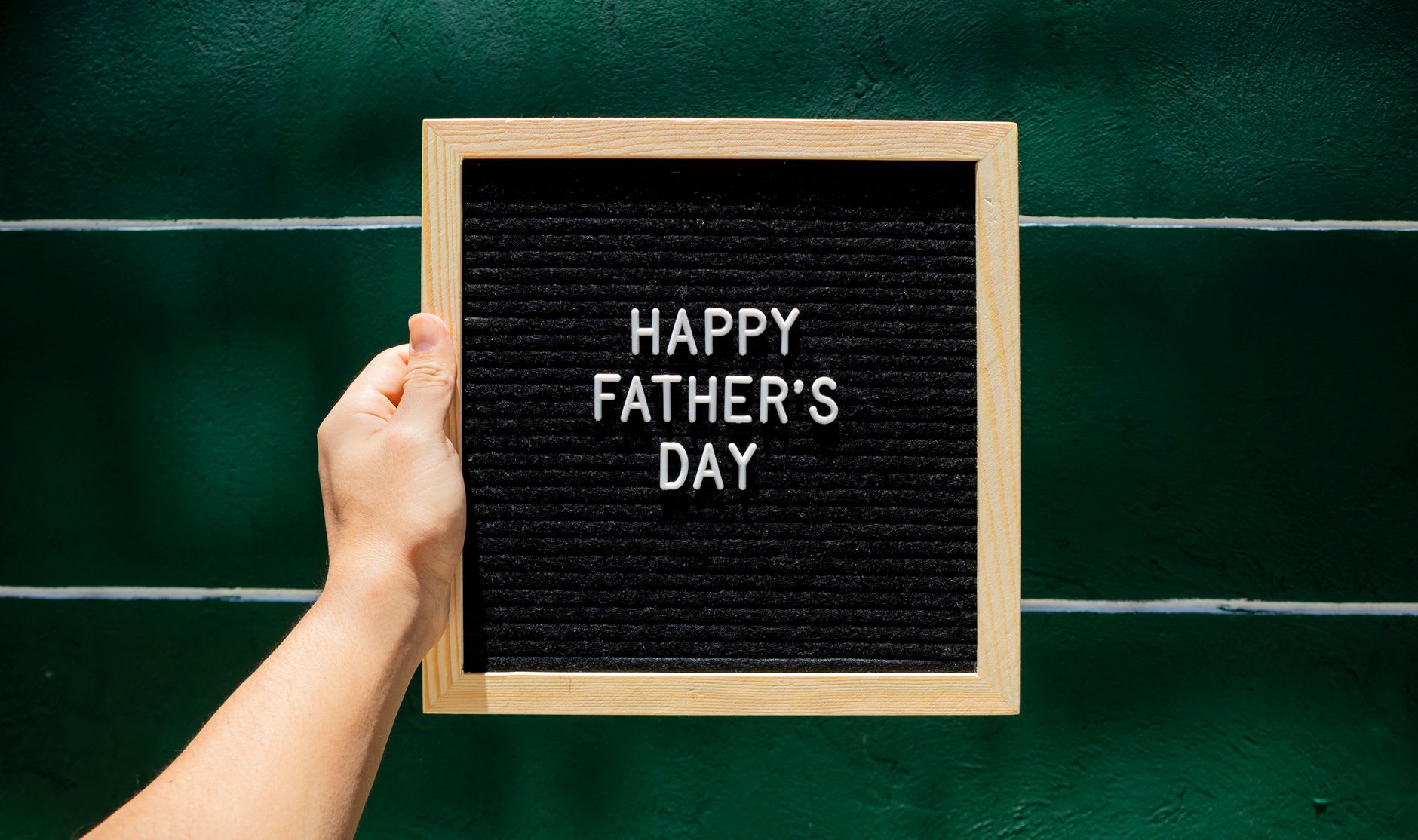 A person holds a glowing 'Happy Father's Day' neon sign against a green wall.