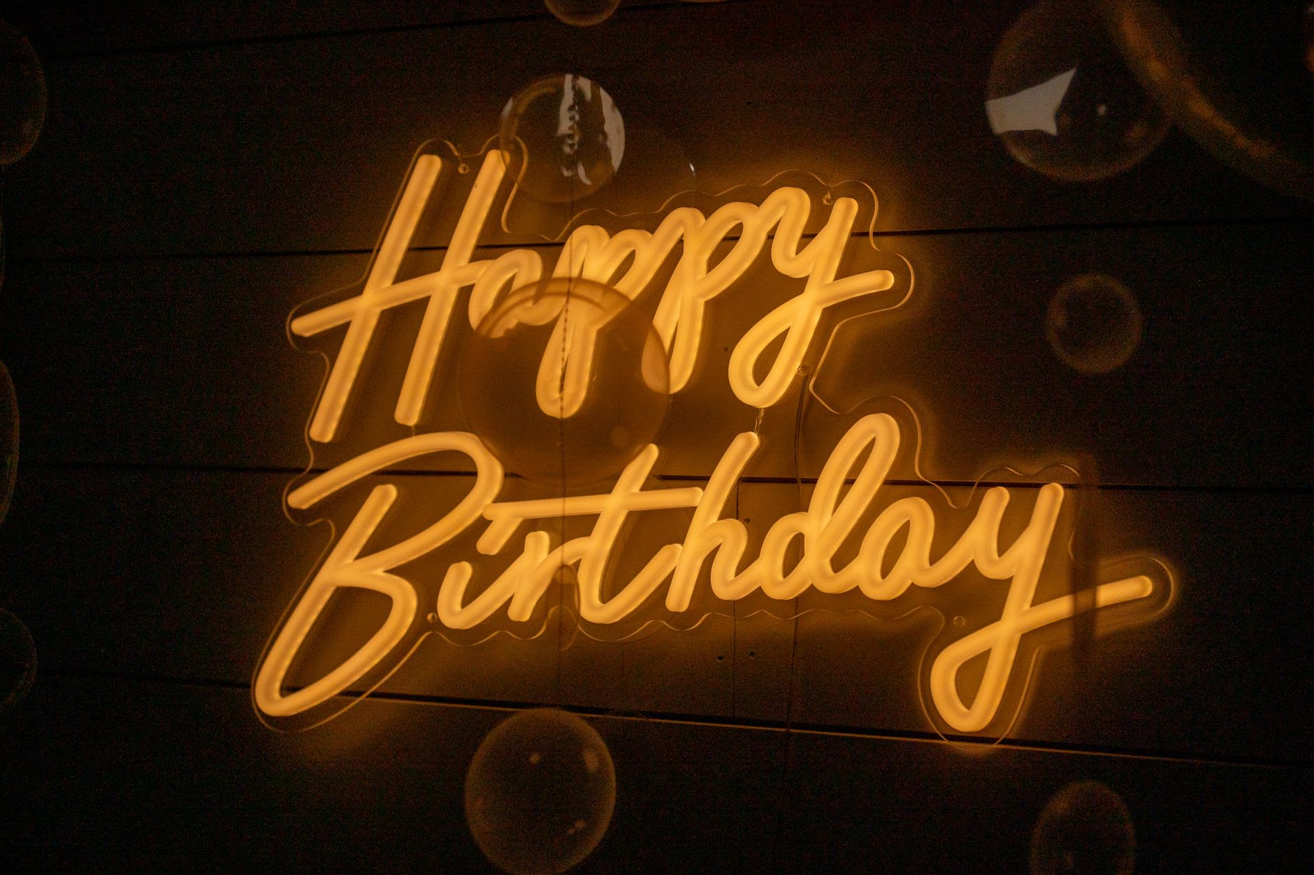 A 'Happy Birthday' neon sign glowing in bright script beside luminous bubble shapes.