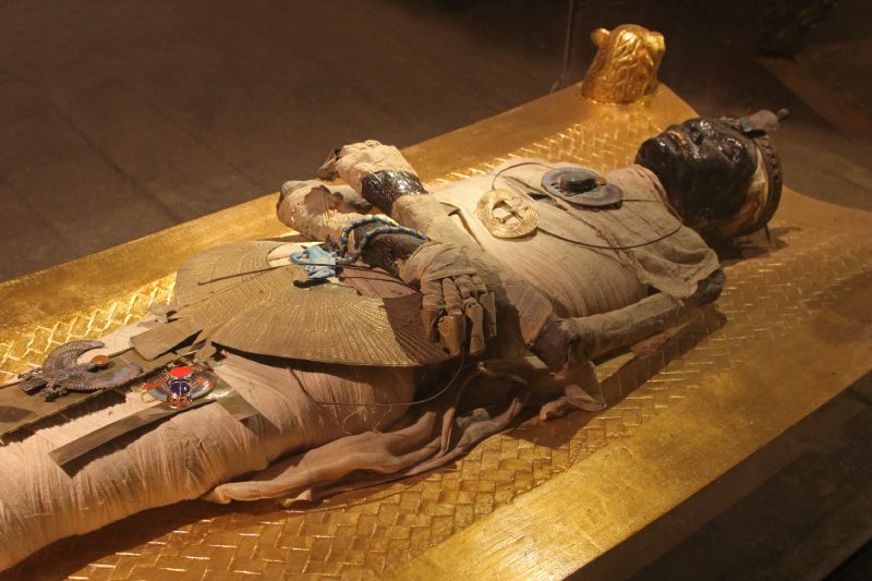 Momia egipcia decorada con amuletos sobre un sarcófago dorado.