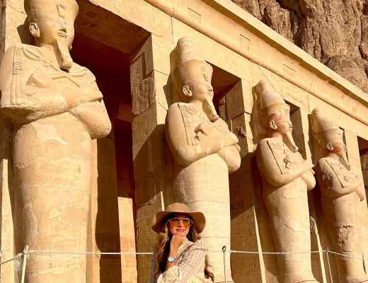 Traveler in cream outfit and wide-brimmed hat seated before monumental ancient Egyptian statues carved into a temple facade.