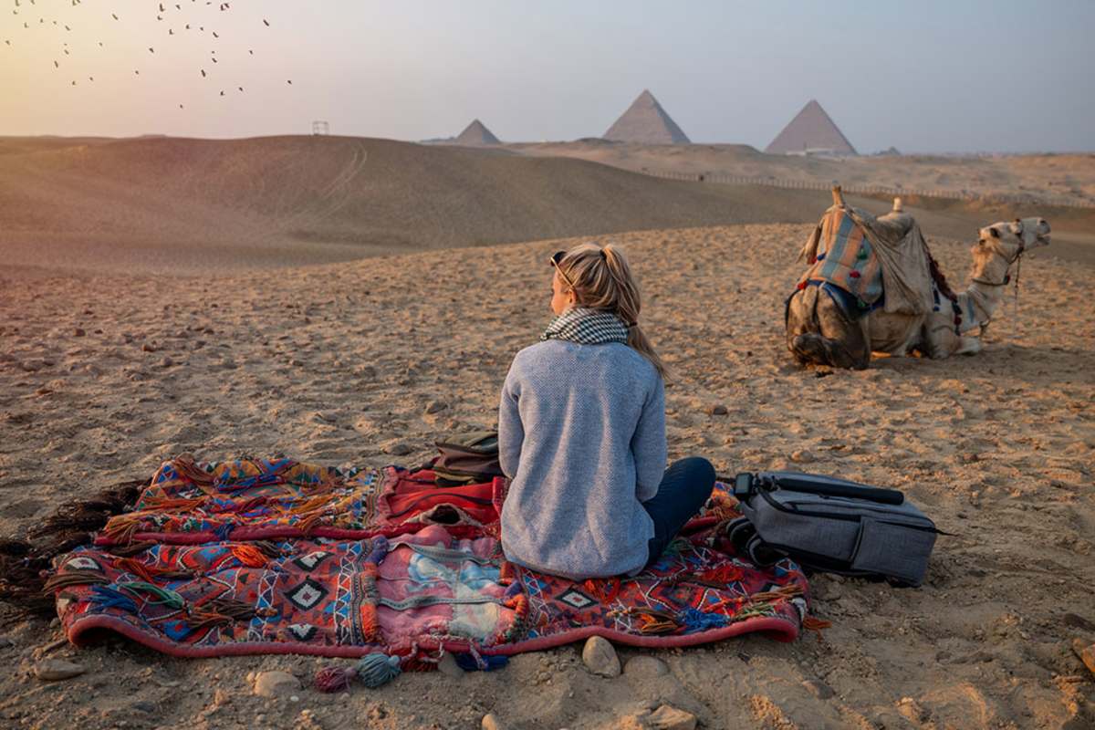a blond girl sitting infront of pyramids of Giza and a camel beside her