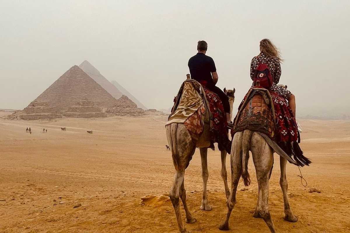 Two travelers riding camels toward the Pyramids of Giza under a hazy desert sky.