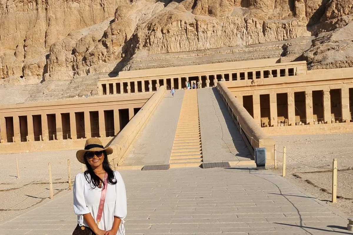 Visitor in white outfit and sunhat standing on pathway leading to the Mortuary Temple of Hatshepsut, built into desert cliffs.