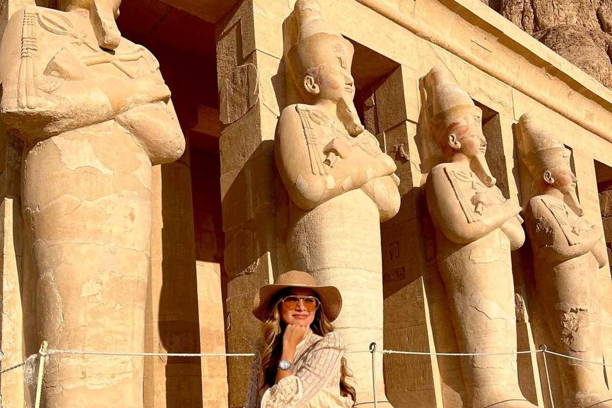 Traveler in cream outfit and wide-brimmed hat seated before monumental ancient Egyptian statues carved into a temple facade.