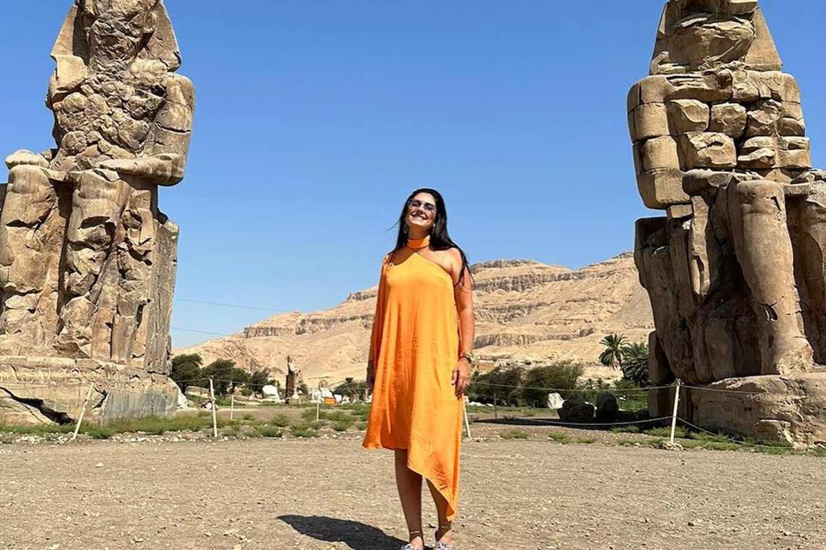 Visitor in bright orange dress standing between the Colossi of Memnon, massive seated stone statues near Luxor under a clear sky.