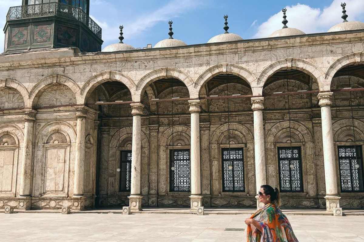 Person in colorful shawl standing in courtyard with ornate arches, domes, and a tall clock tower showing Islamic architectural design.