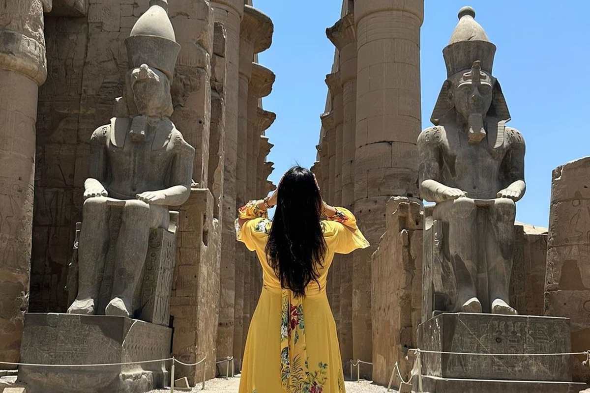 Person in bright yellow floral dress facing massive seated pharaoh statues at temple entrance, with tall stone columns under clear sky.