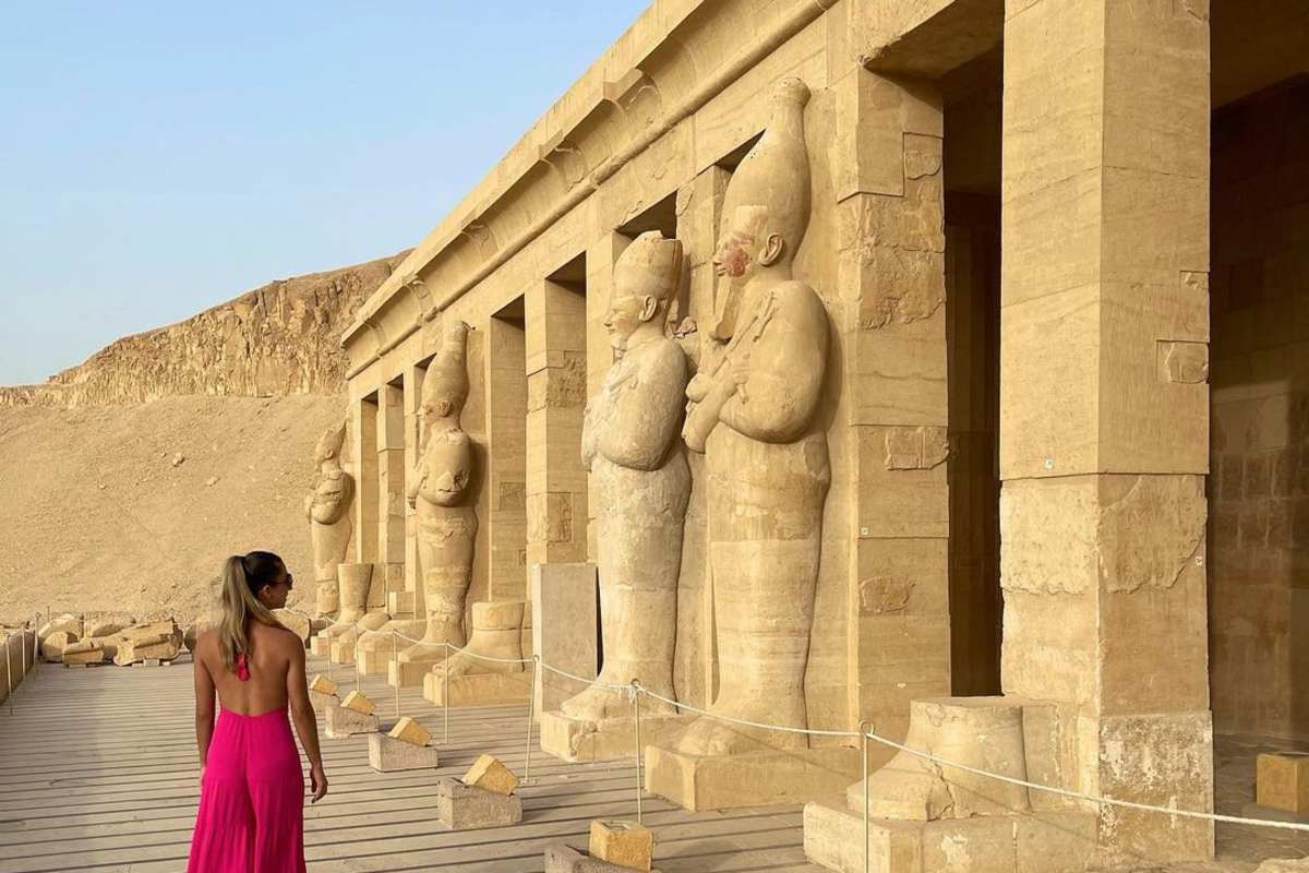 "Person in a bright pink dress walking past colossal ancient statues in an Egyptian temple colonnade.