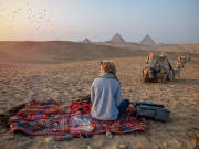 a blond girl sitting infront of pyramids of Giza and a camel beside her