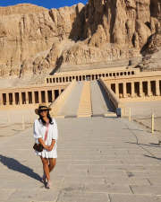 A woman in white dress with a hat stand infront of Hatshepsut temple 