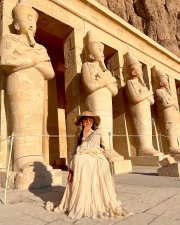 Traveler in cream outfit and wide-brimmed hat seated before monumental ancient Egyptian statues carved into a temple facade.