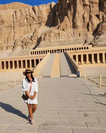 Visitor in white outfit and sunhat standing on pathway leading to the Mortuary Temple of Hatshepsut, built into desert cliffs.