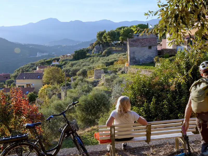 Senior couple talk on bench above forest and villages in autumn, electric bicycles beside them, Liguria
