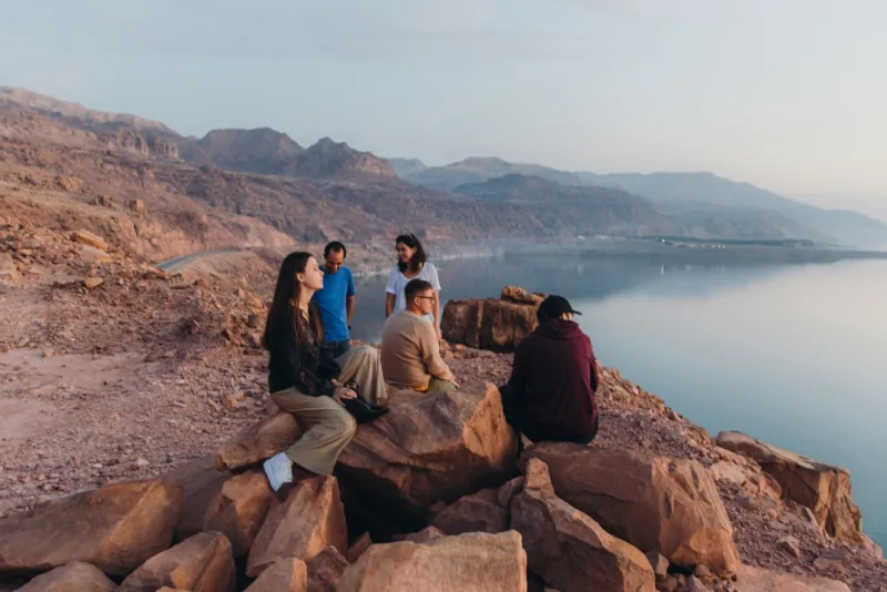 Group of people sitting and standing on rocks overlooking calm lake with rugged mountains in warm light.