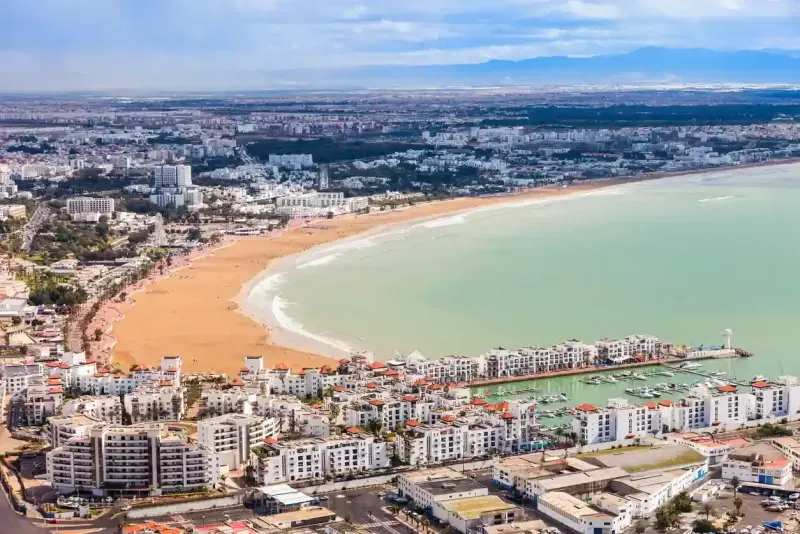 a bright daytime aerial view of Agadir, Morocco, highlighting its golden sandy beach, turquoise bay, and modern whitewashed buildings along the lively waterfront.
