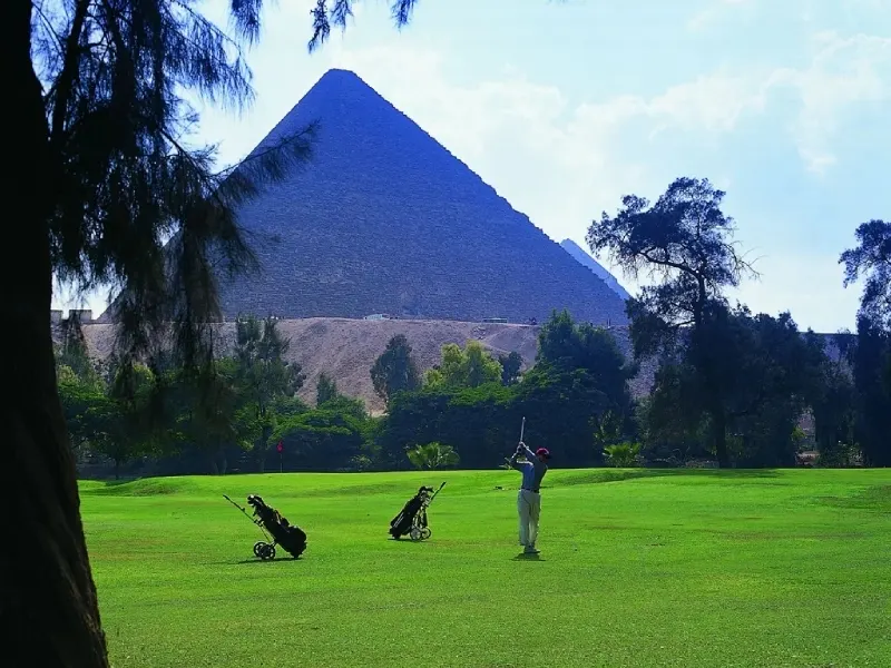 A golfer taking a swing on a green fairway with the Great Pyramid of Giza towering in the background at the Mena House Oberoi Golf Club.
