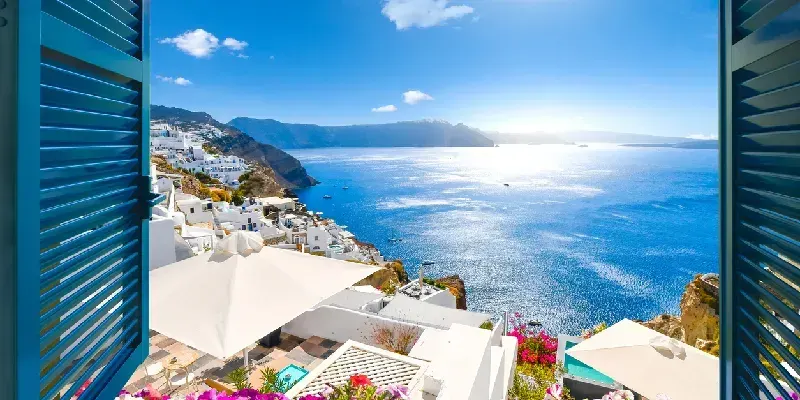 Vista desde una ventana abierta con contraventanas azules del mar Egeo, la caldera, la costa y el pueblo encalado de Oia, Santorini, Grecia.