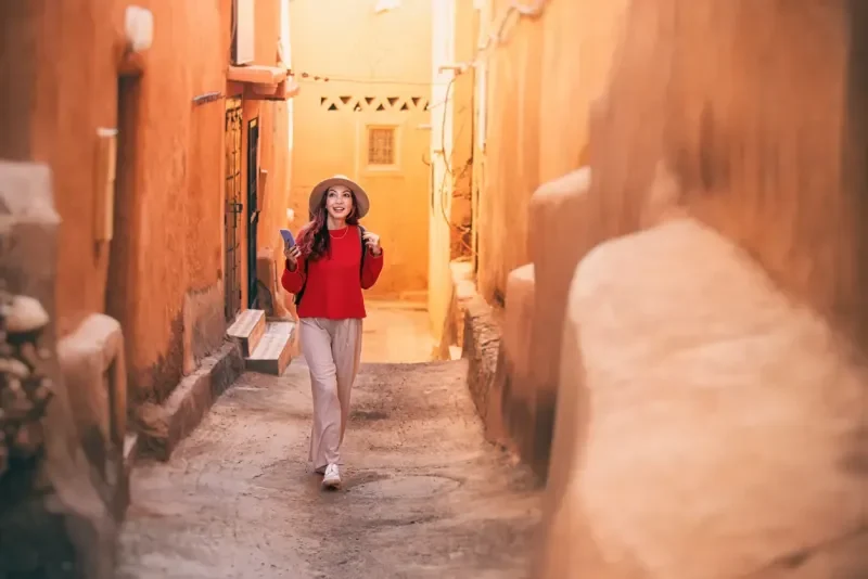 Young woman tourist with backpack and hat is walking in narrow street of Ait Benhaddou, Morocco