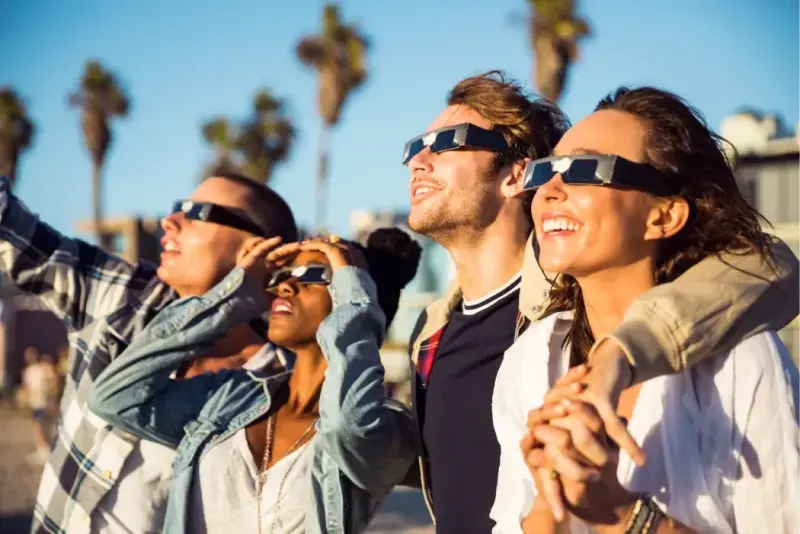L'immagine mostra un gruppo di amici sorridenti che indossano appositi occhiali protettivi per osservare un'eclissi solare.