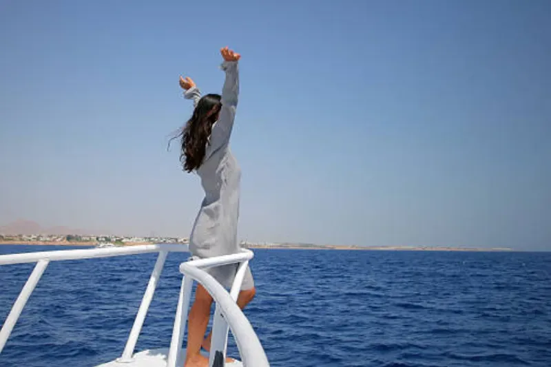 Eine Frau steht am Bug eines weißen Bootes, hebt die Arme in die Luft und genießt den Moment vor einer ruhigen Küstenlandschaft mit blauem Himmel und Meer