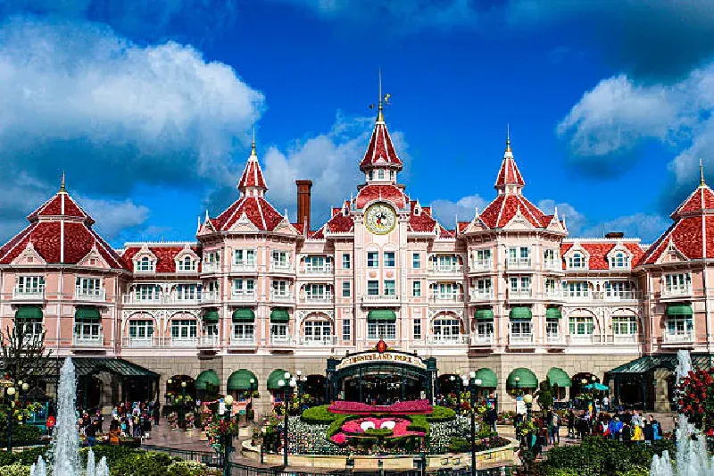 Der Haupt-Eingang von Disneyland mit dem ikonischen Tor, Besucher strömen hinein und bunte Dekorationen im Hintergrund.