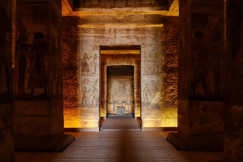 Interior view of Abu Simbel Temple with warm lighting illuminating carved walls and leading to the sanctuary where four seated statues sit in the inner chamber.